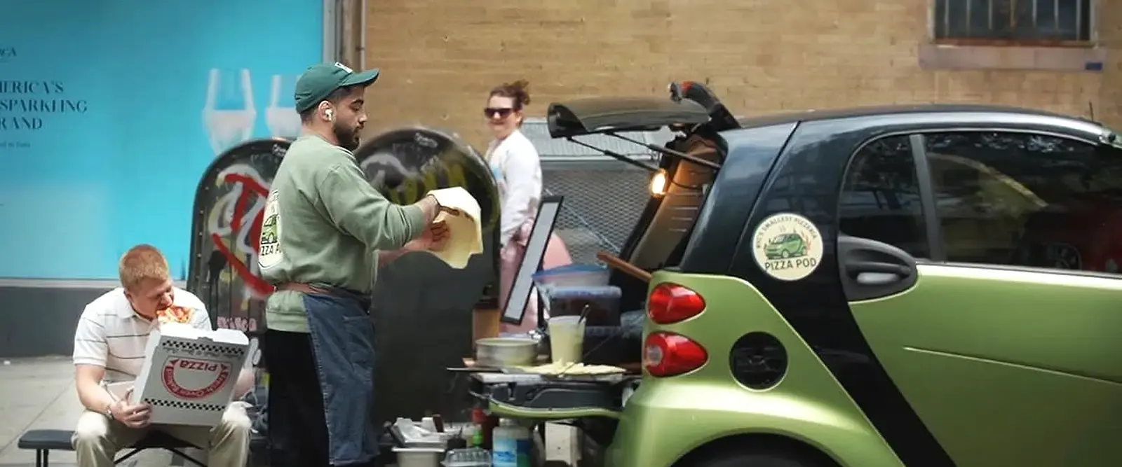 The green Pizza Pod Smart Car parked with its boot open, a cook stretching dough beside it and a customer eating a slice from a box on a bench