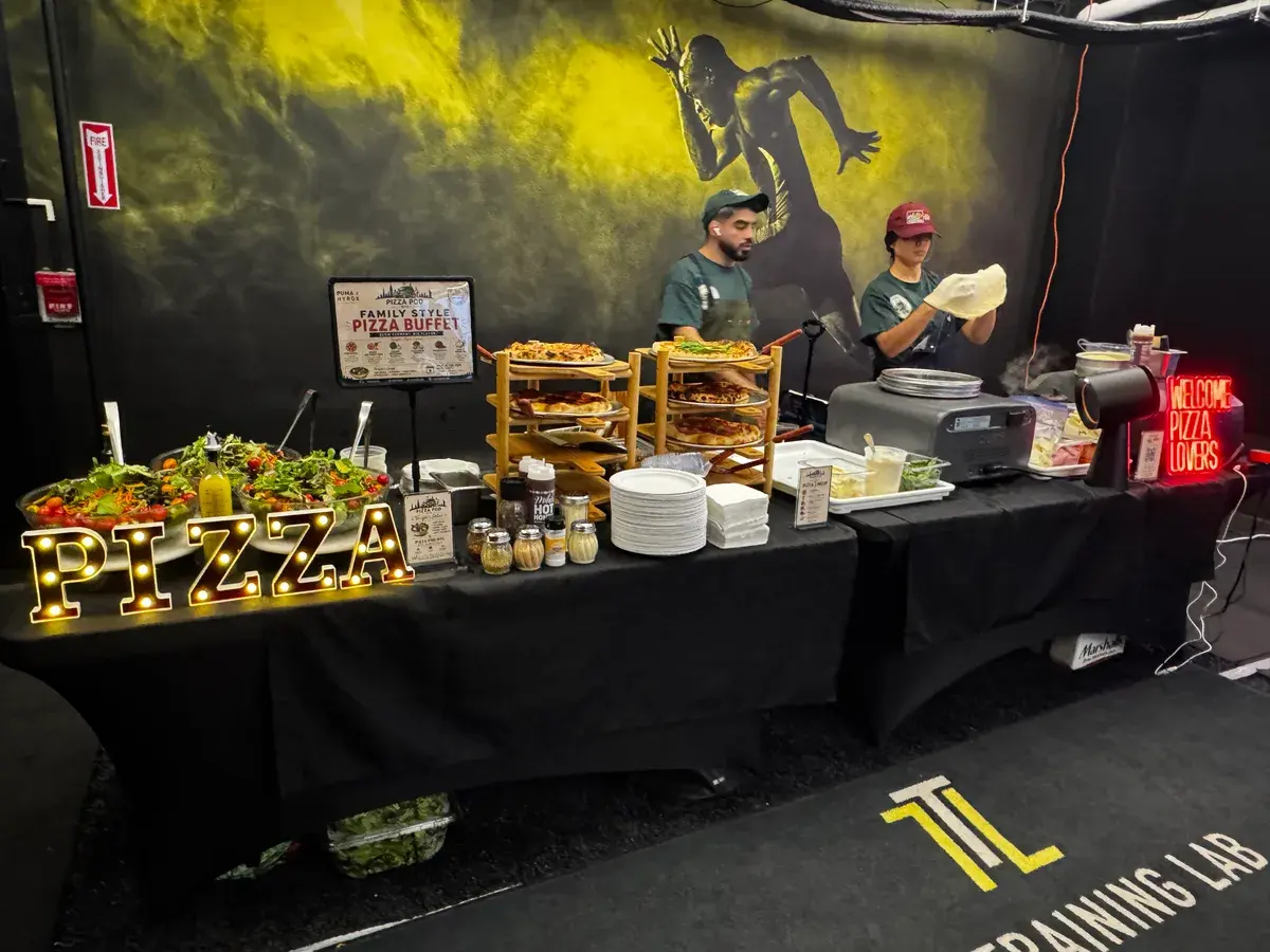 Two Pizza Pod cooks stretching dough behind a buffet table stacked with finished pies